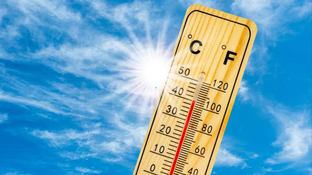 Un termometro di legno sotto un sole splendente e un cielo azzurro con nuvole, che segna una temperatura elevata.