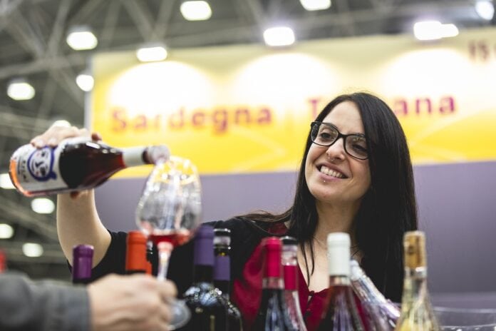 Slow Wine Fair 2026 Una professionista del settore vinicolo sorride mentre versa con cura del vino rosato in un calice di cristallo. Sullo sfondo sfocato si scorgono i cartelli dei padiglioni regionali della Sardegna e della Toscana. In primo piano, una selezione di bottiglie di vino è pronta per la degustazione